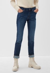 Mörkblå denimjeans med slim fit, uppvikt nederkant, två sidofickor och dragkedjestängning, tillsammans med beiga snörda kängor.
