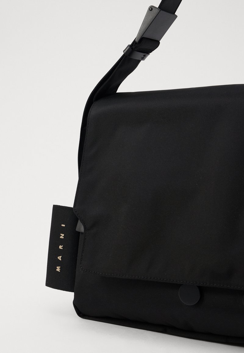 Sac messager en nylon noir avec un rabat repliable, une fermeture à pression et une sangle ajustable. Présente une étiquette logo sur le côté.