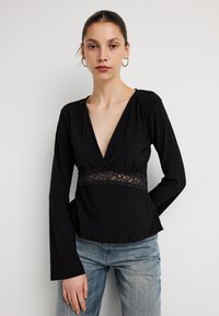 Top negru cu mâneci lungi, decolteu adânc în V și accent din dantelă în talie. Țesătura este netedă, cu o siluetă mulată. Purtați cu blugi albaștri.