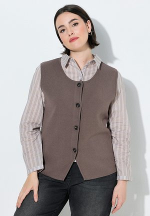Kvinde med kort mørkt hår iført en brun knappet vest over en beige stribet skjorte og mørke jeans, stående mod en hvid væg.