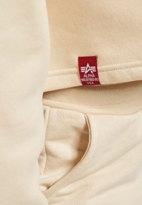 Beige hettegenserstoff med en myk tekstur, med et fremtredende rødt merke som sier "Alpha Industries Inc. U.S.A." nær en lommepart.