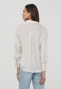 Blusa di seta beige chiaro con uno schienale leggermente trasparente, maniche lunghe, colletto classico e polsini con bottoni, caratterizzata da un design pulito e sartoriale.