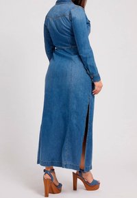 Kvinna klädd i en lång denimklänning med en sidosprund och blå plattformsskor med klumpiga träklackar, stående mot en neutral bakgrund.