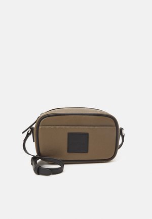 Skórzana torebka crossbody w oliwkowej zieleni z czarną kwadratową naszywką, zamknięciem na zamek i regulowanym paskiem. Gładka faktura, prostokątny kształt.