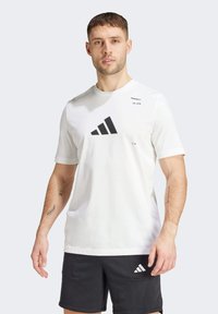 Weißes T-Shirt mit kurzen Ärmeln, das ein großes schwarzes Adidas-Logo und Textakzente zeigt. Aus weichem Stoff gefertigt, mit einem einfachen, sportlichen Design.