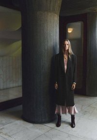 Cămașă lungă neagră peste o rochie roz deschis cu tiv din dantelă, asortată cu cizme maro. Stând lângă un pilon de beton texturat.