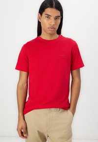 Camiseta de algodón roja con cuello redondo y mangas cortas, que presenta un pequeño logo negro en el pecho. La tela parece suave y ligera.
