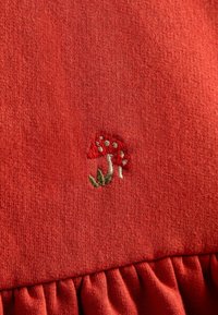 Červená bavlněná tkanina s texturovaným povrchem má malé vyšívané vzory hub v červené, bílé a zelené barvě, ohraničené nařaseným volánem.