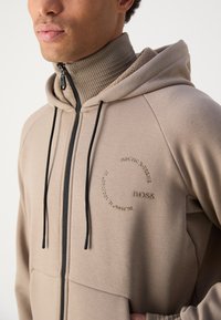 Haut en fleece beige zippé avec un col montant, doté de cordons de serrage noirs et d'un logo circulaire sur la poitrine. La texture semble douce et décontractée.