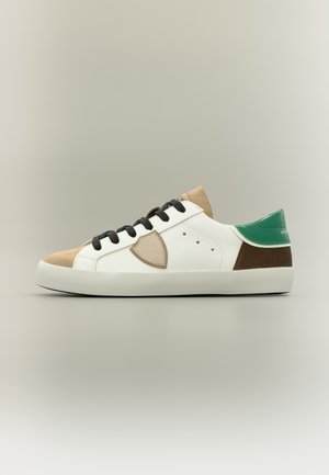 Baskets blanches avec bout beige, lacets noirs, talon vert, détail marron au talon et écusson beige en forme de bouclier sur le côté, sur fond neutre.