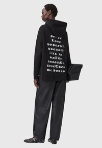 Felpa nera con mantra in argento sul retro, abbinata a pantaloni in pelle nera larghi e ballerine appuntite con borchie, mentre tiene in mano una pochette nera.