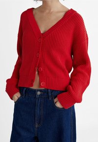 Cardigan rouge en tricot avec un col en V et une texture côtelée, comportant de grands boutons sur le devant et des manches longues et surdimensionnées. Assorti avec un pantalon en denim bleu.