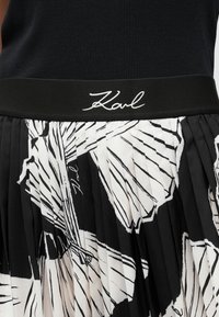 Μαύρη και άσπρη πλισέ φούστα με ανθισμένα σχέδια, με μια σταθερή μαύρη μέση που έχει κεντηθεί με το "Karl" σε λευκή καλλιγραφία.