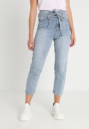 Mujer con jeans cortos de talle alto en azul claro con cinturón anudado y zapatillas blancas, de pie contra un fondo claro y liso.