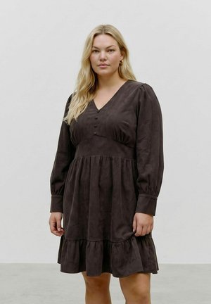 Femme aux longs cheveux blonds portant une robe marron foncé à manches longues avec des boutons et un ourlet à volants, debout devant un fond uni.