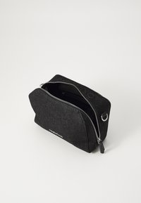 Trousse de maquillage en denim noir avec fermeture éclair. Forme rectangulaire, quincaillerie zipper argentée et branding subtil à l'avant. Compartiments intérieurs visibles.