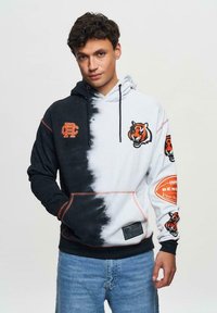 Felpa tie-dye in bianco e nero con dettagli arancioni, dotata di un logo di tigre e grafiche sportive sulle maniche. Realizzata in tessuto morbido con tasca a marsupio.
