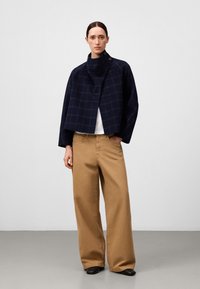 Giacca in lana a scacchi blu navy con colletto alto, abbinata a pantaloni larghi beige. Le scarpe sono nere con design a punta.