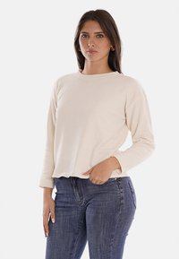 Maglione a maniche lunghe di colore crema con una texture a costine e un collo rotondo, abbinato a jeans di denim scuro. Design semplice e dalla vestibilità rilassata.