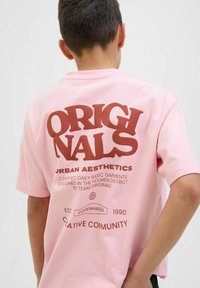Rozā kokvilnas T-krekls ar lielu brūnu tipogrāfiju aizmugurē, ar tekstu "ORIGINALS URBAN AESTHETICS" un papildu dizaina elementiem.