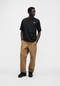 Μαύρο κοντομάνικο μπλουζάκι με μικρό χρυσό λογότυπο στο αριστερό στήθος, συνδυασμένο με χαλαρά παντελόνια χρώματος ταμπά και μαύρα αθλητικά παπούτσια.