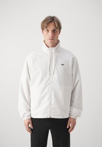 Chaqueta blanca con cremallera y cuello alto, con puños elásticos y un pequeño logo verde de Lacoste en el pecho. Fabricada en un tejido suave y ligero.