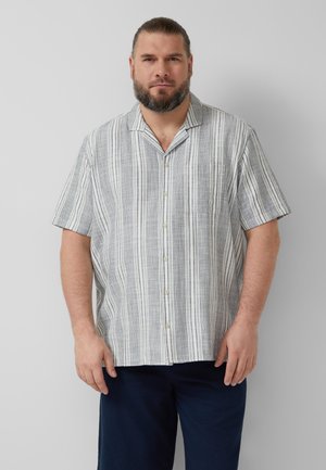 Mężczyzna z brodą, ubrany w koszulę z krótkim rękawem i pionowymi paskami oraz ciemne spodnie, stojący na jednolitym jasnoszarym tle.