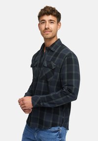 Schwarz-grün kariertes Flanellhemd mit zwei Brusttaschen, Knopfleiste und langen Ärmeln, kombiniert mit hellen Jeans.