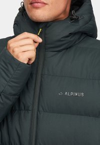 Ciemnozielona kurtka puchowa z wysokim kołnierzem, zapięciem na zamek oraz detalem z logo. Posiada gładką fakturę i elastyczne sznurki do regulacji dopasowania.