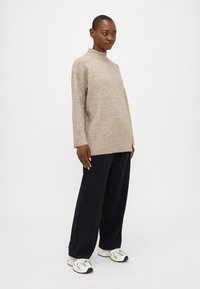 Maglione color marrone chiaro, con collo alto in maglia morbida, presenta maniche lunghe e orlo a costine. Abbinato a pantaloni neri larghi e sneakers bianche.