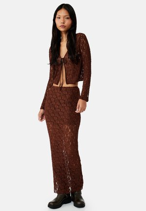 Vrouw staat, draagt een bruin kanten tweedelig set met een lange rok en een lang mouwen top met strik aan de voorkant, gecombineerd met zwarte laarzen.