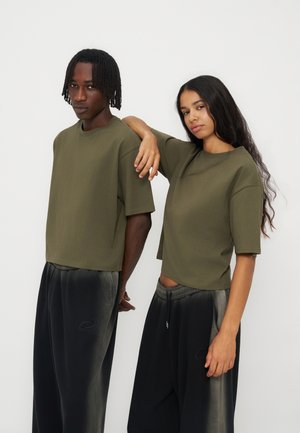 Deux personnes portant des t-shirts vert olive et des pantalons noirs dégradés, debout côte à côte sur un fond clair uni.
