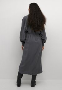 Lång grå klänning med insydd midja, vida ärmar och knäppta manschetter, tillverkad av slät tyg. Bärs med åtsittande svarta byxor.