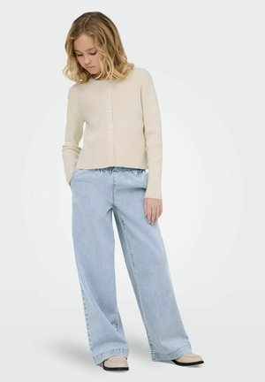 Noor inimene õlgadeni juustega kannab kreemikat nööbitavat kardigani, helesiniseid laia lõikega pükse ja beeže jalanõusid, seisab käed taskutes.