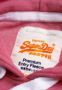 Sudadera con capucha de forro polar rosa con cordones blancos. Presenta una etiqueta con texto naranja: "Vintage SuperDry" y "Premium Entry Fleece".