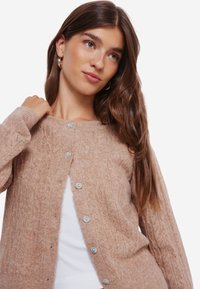 Beige gebreide cardigan met een gestructureerd patroon, voorzien van vijf lichte knopen aan de voorkant, lange mouwen en een ronde halslijn.