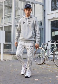 Grå hoodie med "NIKE"-logotyp, vita dragsnören; vita träningbyxor med svarta ränder och logotyp; sportiga skor; urban utomhusmiljö.