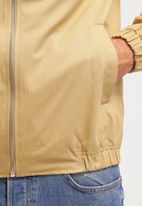 Lyse beige zip-jakke med elastiske manchetter og kant. Glat stof med sidelommer, båret over blå denimjeans. Simpelt og praktisk design.