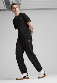 Zwarte T-shirt en joggers van zacht materiaal, met een wit Puma-logo op de borst en het been, gecombineerd met wit-zwarte sneakers.