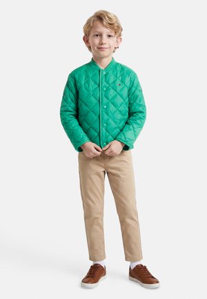 Ξανθό αγόρι όρθιο, φοράει πράσινο μάλλινο μπουφάν, μπεζ παντελόνια, άσπρες κάλτσες και καφέ αθλητικά παπούτσια μπροστά σε λευκό φόντο.