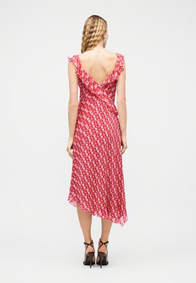 Γυναίκα με κυματιστά ξανθά μαλλιά, ντυμένη με ροζ midi φόρεμα με μοτίβα, χωρίς μανίκια, με βολάν και βαθιά V-πλάτη, και μαύρα ψηλοτάκουνα σανδάλια, όρθια.