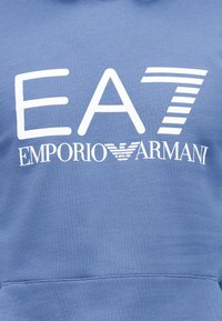Μπλε φούτερ με κουκούλα που διαθέτει έντονο λευκό λογότυπο "EA7" και κείμενο "EMPORIO ARMANI". Κατασκευασμένο από απαλό ύφασμα με βουρτσιστή υφή.