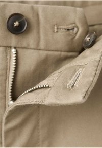 Gros plan de la ceinture d'un pantalon beige montrant une fermeture éclair métallique partiellement ouverte et deux boutons foncés avec des boutonnières.
