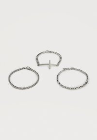 Trois bracelets en chaîne argentée, dont un avec un pendentif croix horizontale, disposés sur un fond blanc.