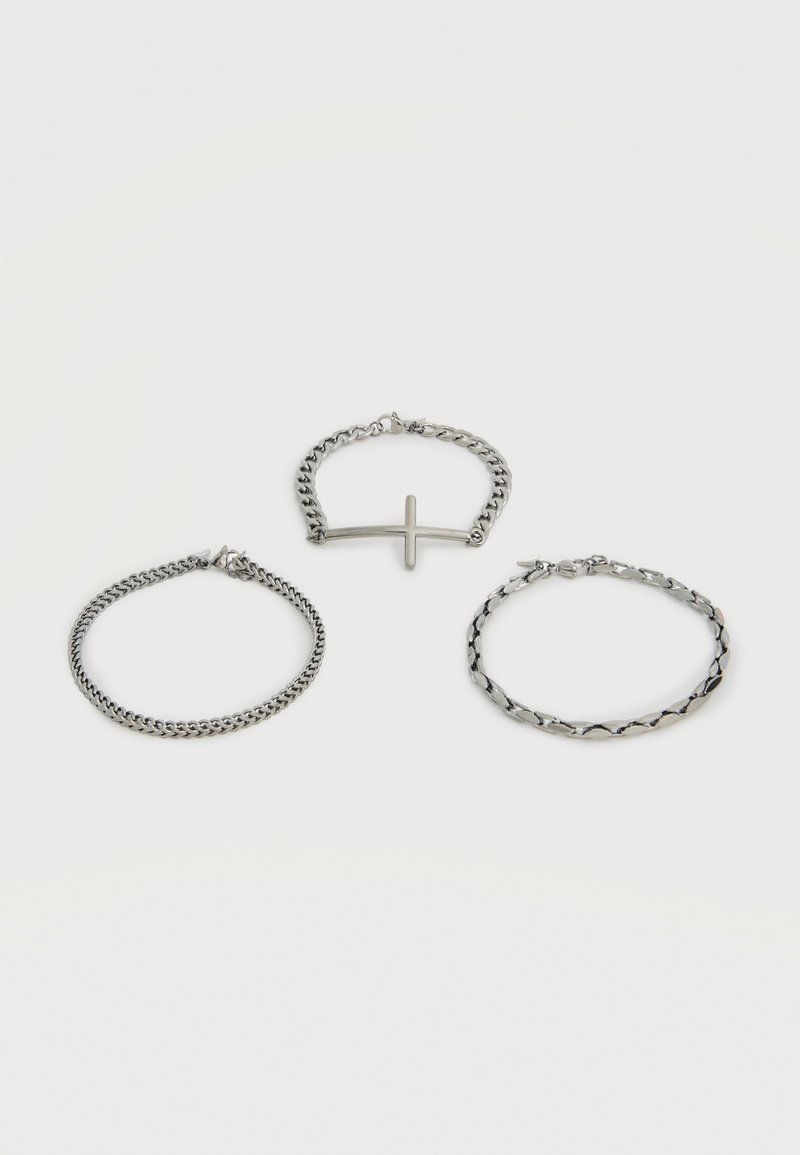 Trois bracelets en chaîne argentée, dont un avec un pendentif croix horizontale, disposés sur un fond blanc.