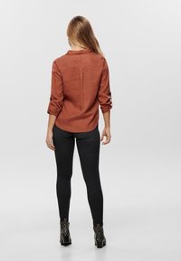 Eisenrostfarbendes, langärmliges Hemd mit hochgerollten Ärmeln, runder Saum und detailierter Rückenseite, kombiniert mit schwarzen Skinny-Jeans und Schlangenlederprint-Stiefeletten.