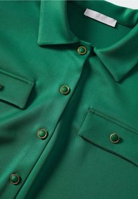 Zielona satynowa bluzka z kołnierzykiem, dwiema kieszeniami na piersi i złotym wykończeniem. Gładka tekstura, z zaokrąglonymi detalami przy guzikach.