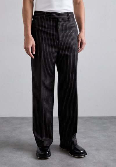 Pantalon noir à rayures avec une coupe ajustée, présentant une texture lisse et un devant plat, associé à des chaussures noires.