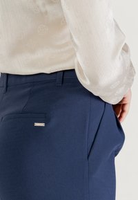 Pantalon bleu marine à texture lisse, doté d'une poche arrière unique et d'une petite étiquette de marque en argent. Associé à un chemisier léger à motifs.