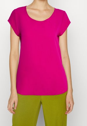 Femme portant un chemisier rose vif à manches courtes avec un col rond et un pantalon vert olive, debout devant un fond blanc.